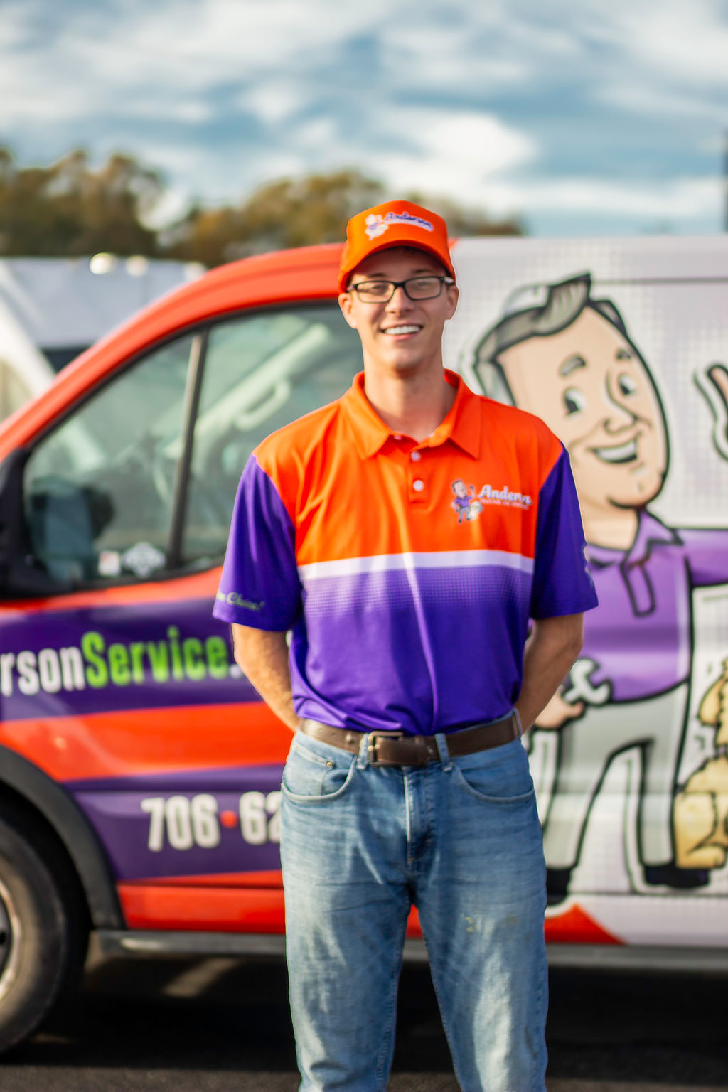 An Anderson Heating, Air & Insulation team member smiling in a branded Anderson Heating, Air & Insulation polo and hat in front of a fully wrapped Anderson service van at the Calhoun headquarters — the kind of family-culture, family-owned team Anderson hires.