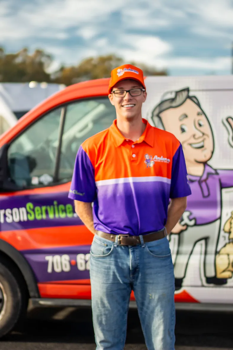 An Anderson Heating, Air & Insulation team member smiling in a branded Anderson Heating, Air & Insulation polo and hat in front of a fully wrapped Anderson service van at the Calhoun headquarters — the kind of family-culture, family-owned team Anderson hires.